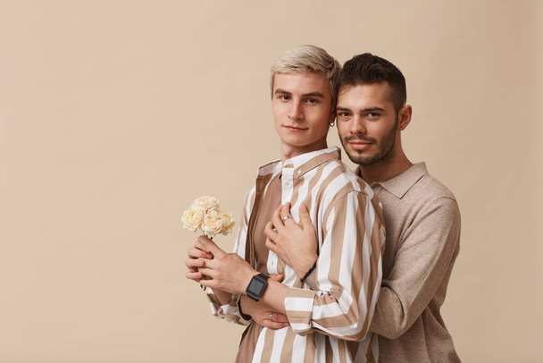 Ritratto minimale in vita di una giovane coppia gay che abbraccia e guarda la fotocamera mentre posa con fiori sullo sfondo beige neutro in studio, copia lo spazio - Foto, immagini