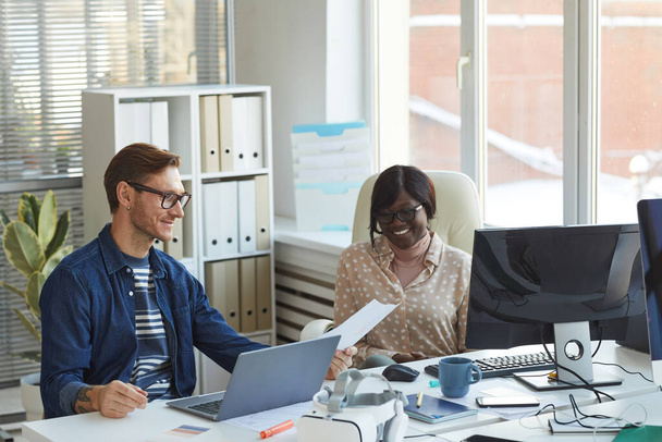 Retrato de um empresário contemporâneo sorridente conversando com colega afro-americano enquanto usa computadores no interior do escritório branco - Foto, Imagem