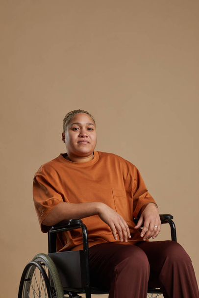 Verticaal portret van hedendaagse gemengde ras vrouw met behulp van een rolstoel en kijken naar de camera terwijl poseren in de studio tegen een neutrale achtergrond - Foto, afbeelding