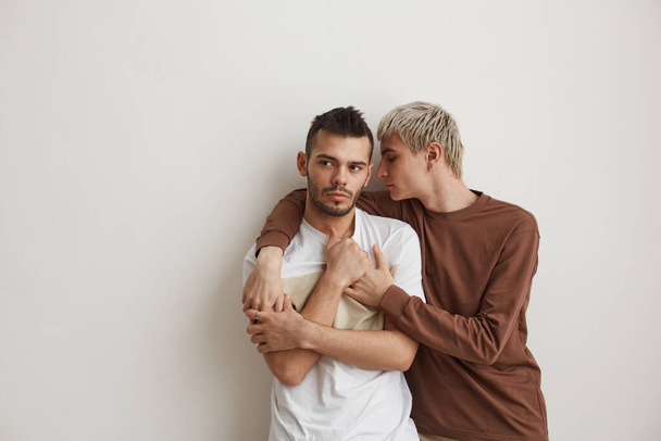 Cintura mínima hasta el retrato de la joven pareja gay abrazándose mientras está de pie contra la pared blanca en el interior, espacio de copia - Foto, imagen