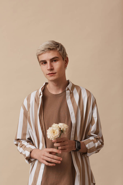 Vertical portrait of feminine blonde man holding flowers and looking at camera while standing against neutral beige background in studio - Fotografie, Obrázek