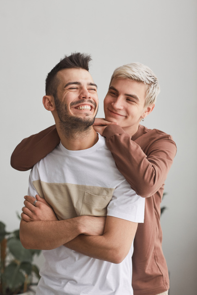 Otevřený portrét bezstarostného homosexuálního páru objímajícího se uvnitř a šťastně se smějícího při pózování proti bílému - Fotografie, Obrázek