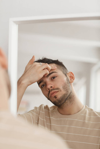 Portrait vertical du jeune homme inspectant sa peau tout en regardant dans le miroir pendant la routine du matin, espace de copie - Photo, image