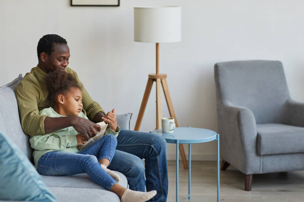 Portrait of loving African-American father and daughter sitting on couch together in cozy home interior, copy space - Foto, imagen