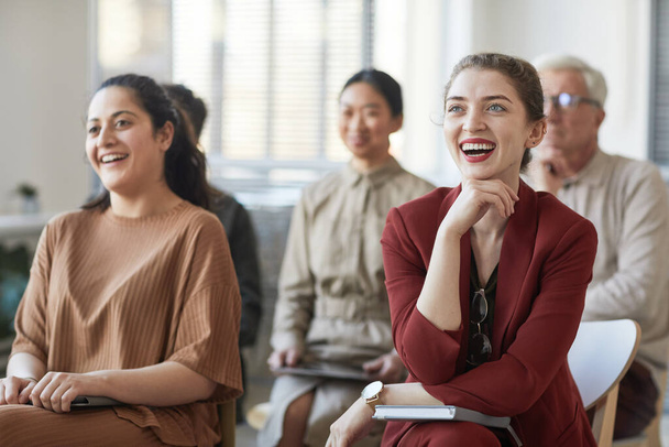 Разнообразная группа деловых людей, сидящих на стульях в аудитории и слушающих на совещании или семинаре, фокусируется на двух улыбающихся деловых женщинах на переднем плане - Фото, изображение