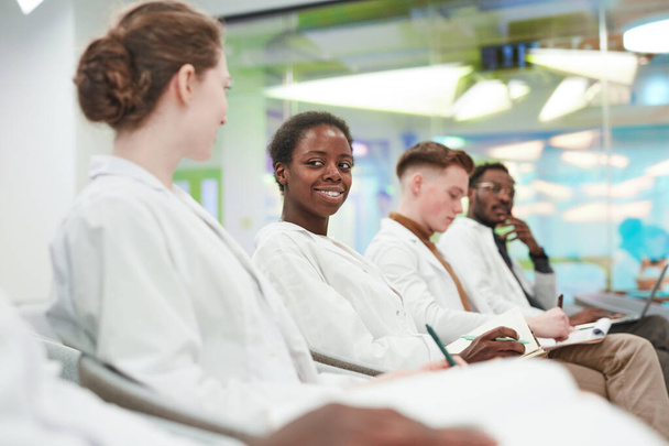 Oldalnézet a többnemzetiségű fiatalok csoportjában, akik laborköpenyt viselnek, miközben sorban ülnek és orvosi előadást hallgatnak - Fotó, kép