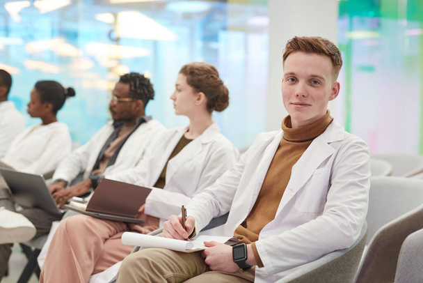 Muotokuva nuori mies yllään lab takki ja hymyilee kameralle istuessaan peräkkäin monikansallisen ryhmän yleisöä lääketieteellisessä seminaarissa, kopioi tilaa - Valokuva, kuva