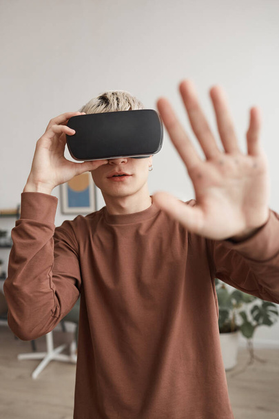 Retrato vertical de la cintura hacia arriba del joven rubio usando VR y extendiendo la mano en el interior del hogar - Foto, Imagen