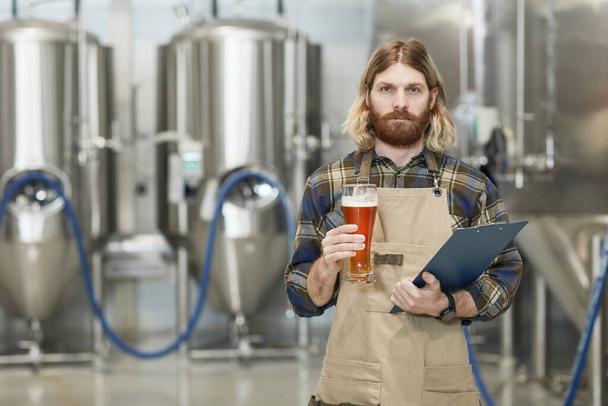 ビールグラスを持ち、醸造工場でワークショップに立っている間にカメラを見ているひげを生やした醸造長の腰の肖像画、コピースペース - 写真・画像