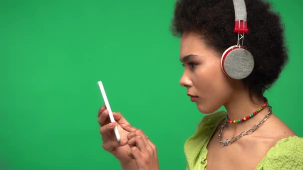 Αφροαμερικανή γυναίκα με ακουστικά και κινητό να τραγουδά απομονωμένη στο πράσινο  - Πλάνα, βίντεο