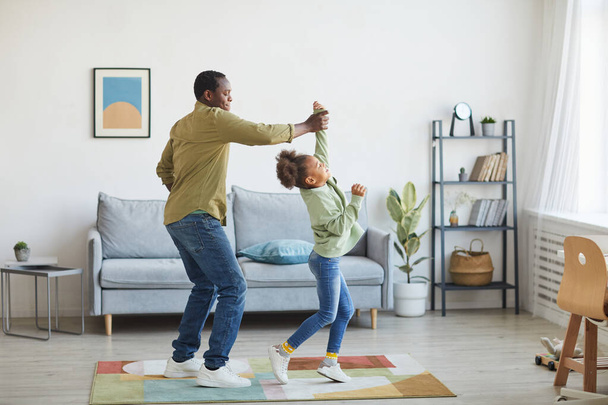 Повний портрет щасливого афро-американського батька, який танцює з дочкою, насолоджуючись мінімальним внутрішнім простором. - Фото, зображення