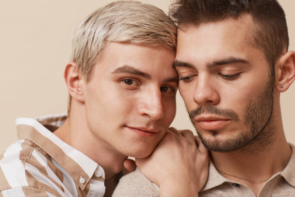 Close up portrait of happy gay couple looking at camera while posing against beige background in studio - Photo, Image