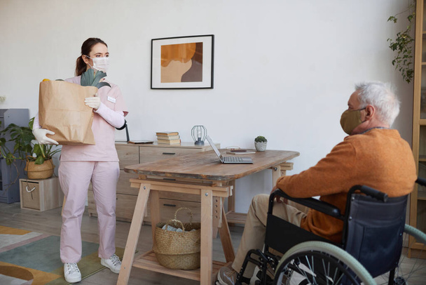 Full length portrait of young woman holding bag with groceries while delivering food to senior man in wheelchair, copy space - Zdjęcie, obraz