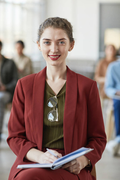 Vertikales Porträt einer erfolgreichen jungen Geschäftsfrau, die rot trägt und in die Kamera lächelt, während sie im Büro auf einem Stuhl sitzt - Foto, Bild