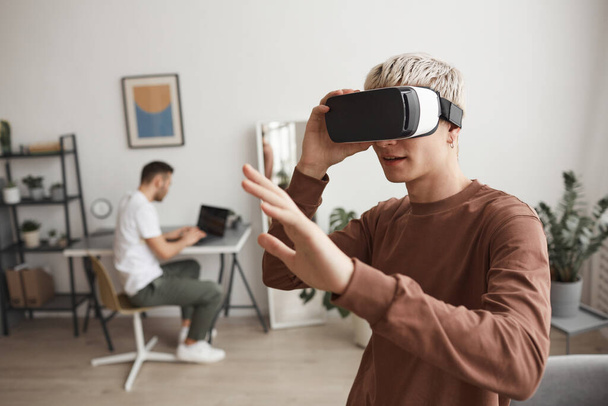 Σηκώστε το πορτρέτο του ξανθού νεαρού άνδρα που χρησιμοποιεί VR και απλώστε το χέρι σας στο εσωτερικό του σπιτιού, αντιγράψτε χώρο - Φωτογραφία, εικόνα
