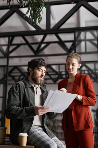 Vertical portrait of contemporary bearded businessman talking to female colleague and holding documents in graphic office interior - Photo, Image