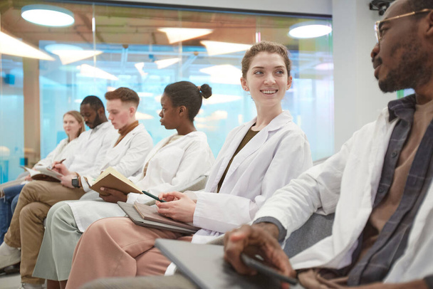 Wieloetniczna grupa młodych ludzi noszących fartuchy laboratoryjne i uśmiechających się siedząc w rzędzie i podczas wykładu na temat medycyny w koledżu, kopiuj przestrzeń - Zdjęcie, obraz