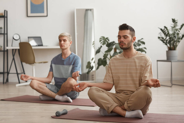 Полный портрет двух молодых людей, медитирующих дома, наслаждаясь упражнениями йоги, сидя на ковриках в позе лотоса - Фото, изображение