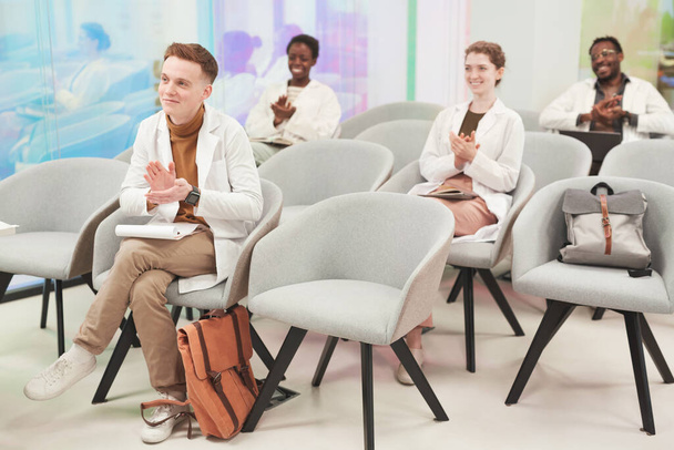 Täyspitkä muotokuva monikansallisesta ihmisryhmästä, joka taputtaa istuessaan yleisössä ja kuunnellessaan luentoa lääketieteestä yliopistossa tai coworking-keskuksessa, kopioi tilaa - Valokuva, kuva