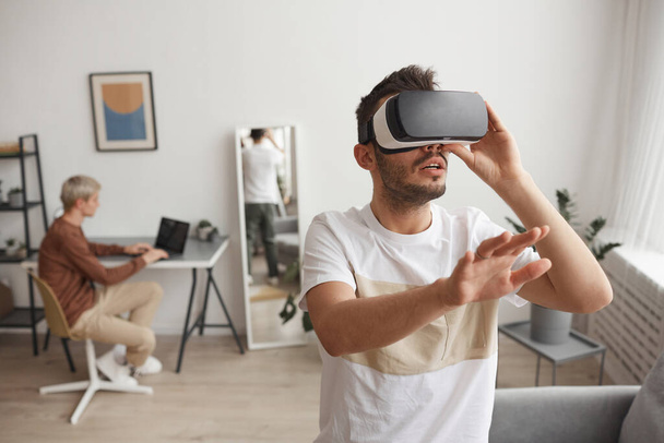 Retrato de la cintura hacia arriba del hombre joven usando VR y extendiendo la mano en el interior del hogar, espacio de copia - Foto, imagen