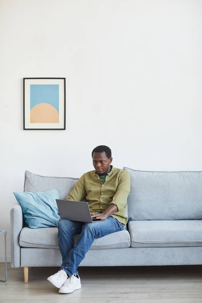 Вертикальный портрет взрослого афроамериканца в полный рост с ноутбуком, сидя на диване в минимальном домашнем интерьере, пространство для копирования - Фото, изображение