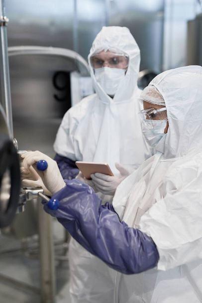 Vista lateral a dos trabajadores que usan trajes de protección mientras usan máquinas en plantas químicas modernas - Foto, Imagen