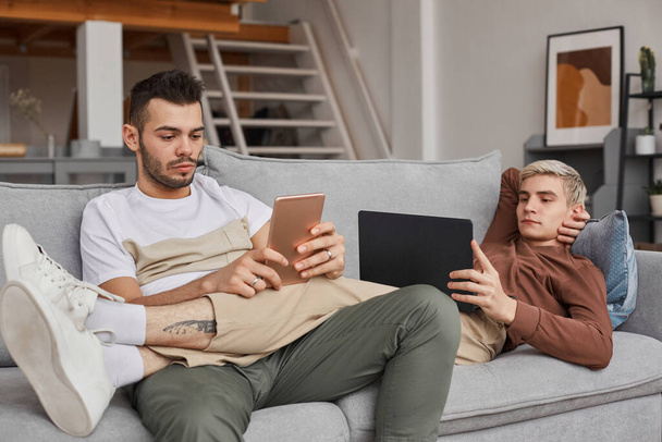Portré kortárs meleg pár használ számítógépet, miközben pihennek a kanapén együtt minimális otthoni belső, másolás tér - Fotó, kép