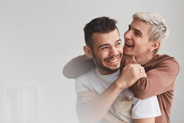 Candid cintura arriba retrato de despreocupada pareja gay abrazando en el interior y riendo felizmente mientras posando contra blanco, espacio de copia - Foto, Imagen