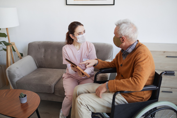 Nagylátószögű portré fiatal női ápolónő asszisztál idős férfi kerekesszékes segítségével digitális tablet az idősek otthonában, mindkettő visel maszk, másolás tér - Fotó, kép