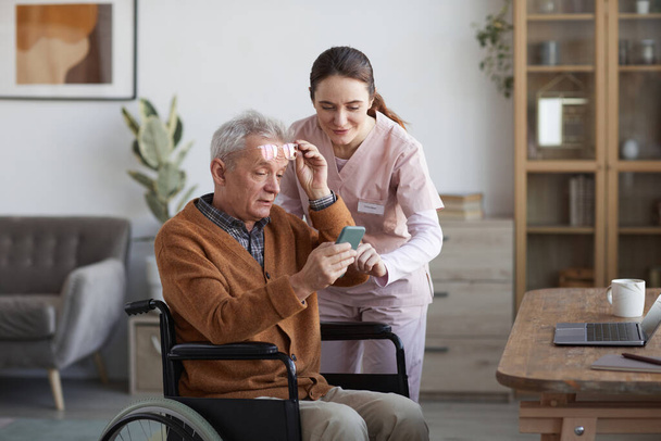 Idősebb kerekesszékes férfi portréja okostelefonnal és gondozóval, aki segít neki az idősek otthonában, másolja a helyet - Fotó, kép