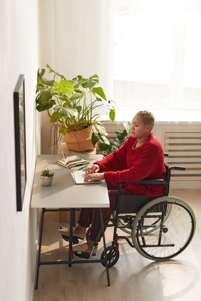 Portrait vertical complet de la femme mixte moderne utilisant un fauteuil roulant tout en travaillant de la maison au bureau éclairé par la lumière du soleil, espace de copie - Photo, image