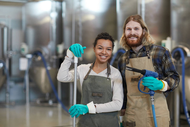 Waist up portrait of two smiling young people wearing aprons and looking at camera during cleaning at industrial factory, copy space - Photo, Image