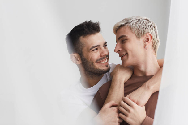Candid κλείσει πορτρέτο του νεαρού gay ζευγάρι αγκαλιάζει τρυφερά και χαμογελώντας, ενώ στέκεται από το παράθυρο στο σπίτι κατά λευκό, αντίγραφο χώρο - Φωτογραφία, εικόνα