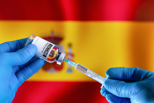 Doctor preparing vial of vaccine injection for the vaccination plan against diseases in Spain. Preparing dose of vaccine in syringe for infections prevention in front of the Spanish flag - Photo, Image