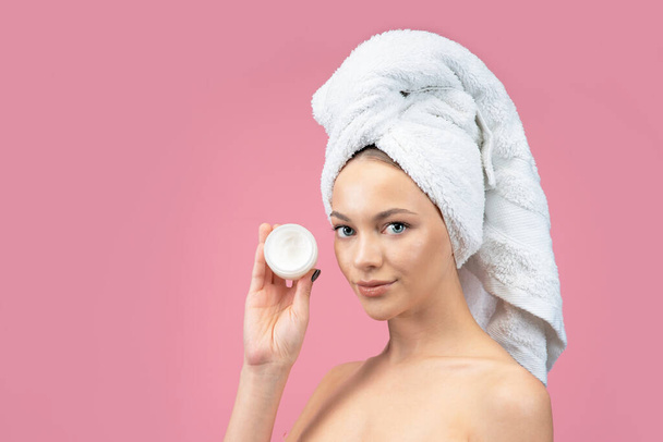 A young, beautiful, fresh beauty woman holds a jar of cream in her hands. female with towel on her head after spa, radiant face and skin care. On a monochrome pink background - Valokuva, kuva