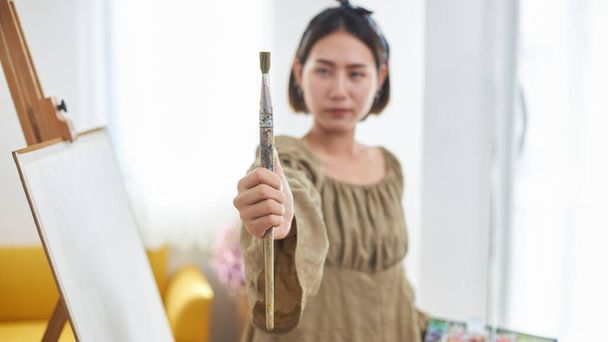 Influenciador creativo o blogger mujer una demostración de pintura en streaming en línea. Una joven artista asiática sosteniendo una paleta de pintura y pinceles está transmitiendo en vivo con un teléfono inteligente en casa. - Foto, imagen