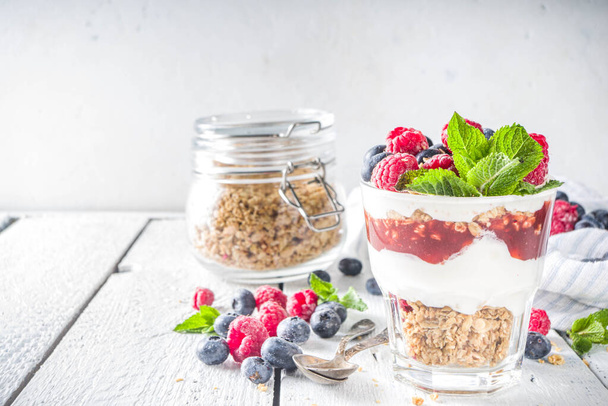 Yogurt parfafait with granola and berries. Sweet and healhty breakfast dessert in glass with granola, Yogurt, blueberries and raspberries. White wooden background top view - Foto, Imagem