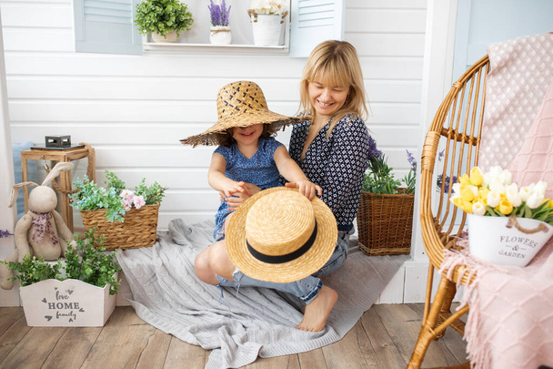 little girl of 4-5 years old is playing with straw hats on veranda of house with mother in her arms. Laughter, happiness, joy, motherhood concept - Foto, imagen