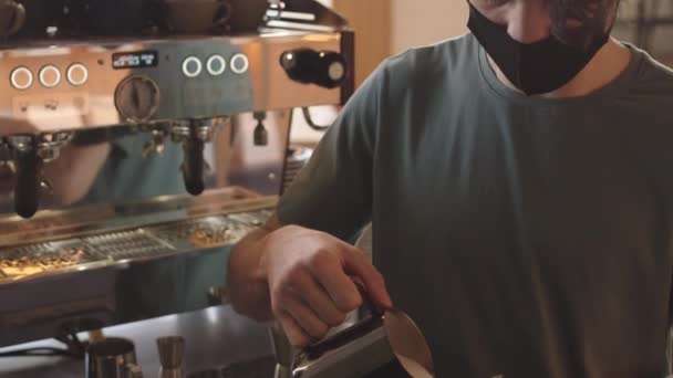 Наклон молодой бармен в футболке и маске стоя в баре рядом с автоматической кофеваркой с чашкой и кувшином в руках и сделать капучино - Кадры, видео