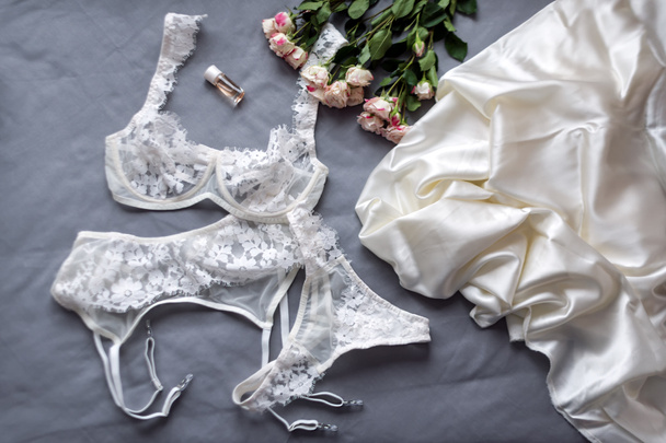 Set of glamorous stylish sexy lace lingerie with woman accessories and flowers on grey bedsheets background. Flat lay woman essentials for holiday. Shopping and fashion concept. - Фото, изображение