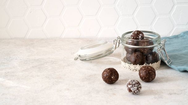 Healthy energy protein balls with nuts, dates, peanut butter, cocoa powder, coconut flakes, oats in glass jar on the kitchen. Homemade truffles. Vegan raw fitness food snack. Side view. Copy space. - Fotografie, Obrázek