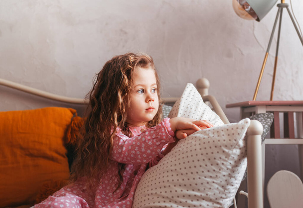 A little sad girl sits on a bed in the bedroom and looks out the window. children's emotions. Melancholic mood - Foto, Imagen