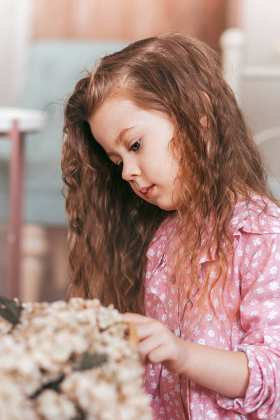Ένα μικρό όμορφο κορίτσι με μαλλιά Fornic κοιτάζει τα λουλούδια. Κάθετη απεικόνιση - Φωτογραφία, εικόνα
