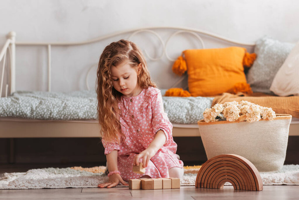 Ένα μικρό κορίτσι παίζει με ξύλινους κύβους στο πάτωμα στο παιδικό δωμάτιο - Φωτογραφία, εικόνα