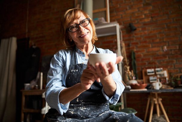 Nice female potter demonstrating clay bowl in pottery workshop - Photo, image