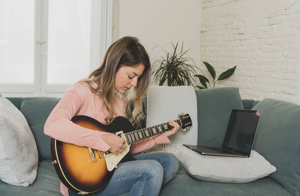 Attractive musician woman playing electric guitar during online concert at home in isolation. Music teacher teaching playing instrument online using laptop, streaming and recording courses. - 写真・画像