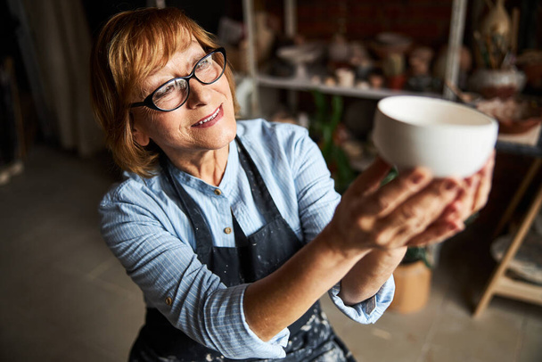 Charmant potier féminin regardant bol d'argile dans l'atelier de poterie - Photo, image