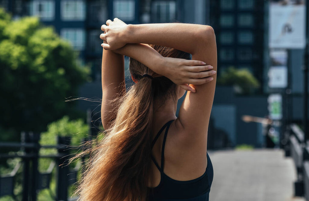 Οπίσθια όψη γυναίκας που τεντώνει το χέρι και τον ώμο της έξω στην οδό Σίτυ. Κοντινή φωτογραφία γυμναστικής με αθλητικά που απλώνει τα χέρια της στην πόλη - Φωτογραφία, εικόνα