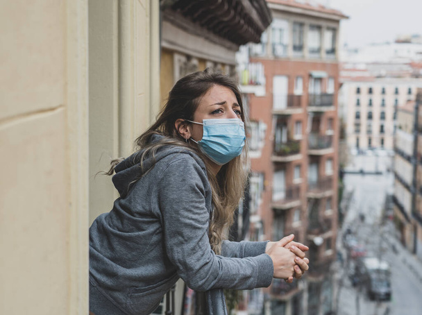 Beautiful depressed lonely woman with face mask in quarantine staring out on balcony feeling sad, lonely, pain and grief during coronavirus outbreak. Depression and mental health due to COVID-19. - Фото, изображение
