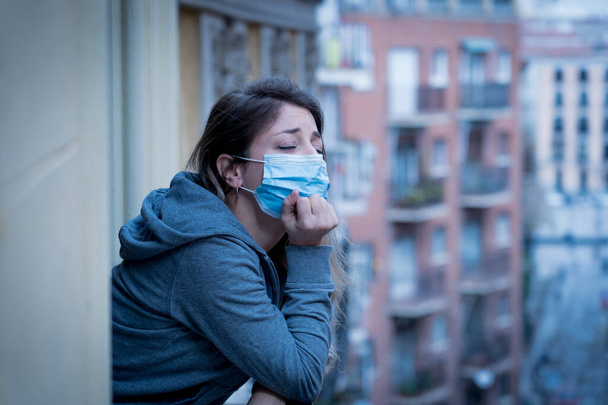 Beautiful depressed lonely woman with face mask in quarantine staring out on balcony feeling sad, lonely, pain and grief during coronavirus outbreak. Depression and mental health due to COVID-19. - Fotoğraf, Görsel